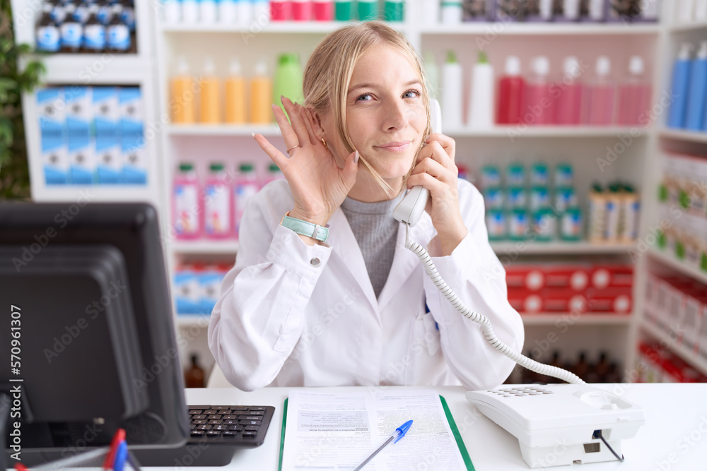 Young caucasian woman working at pharmacy drugstore speaking on the telephone smiling with hand over ear listening an hearing to rumor or gossip. deafness concept. Young caucasian woman working at pharmacy drugstore speaking on the telephone smiling with hand over ear listening an hearing to rumor or gossip. deafness concept.