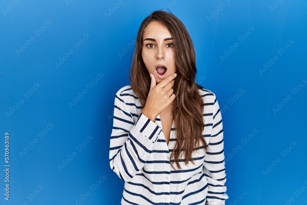 Young hispanic woman standing over blue isolated background looking fascinated with disbelief, surprise and amazed expression with hands on chin Young hispanic woman standing over blue isolated background looking fascinated with disbelief, surprise and amazed expression with hands on chin
