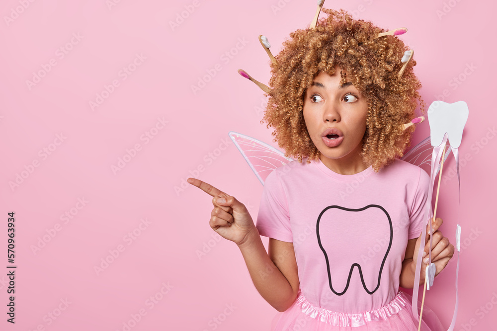 Stunned female model pretends being tooth fairy holds magic wand stares with omg expression points index finger aside reacts to something stunning isolated over pink background copy space for advert