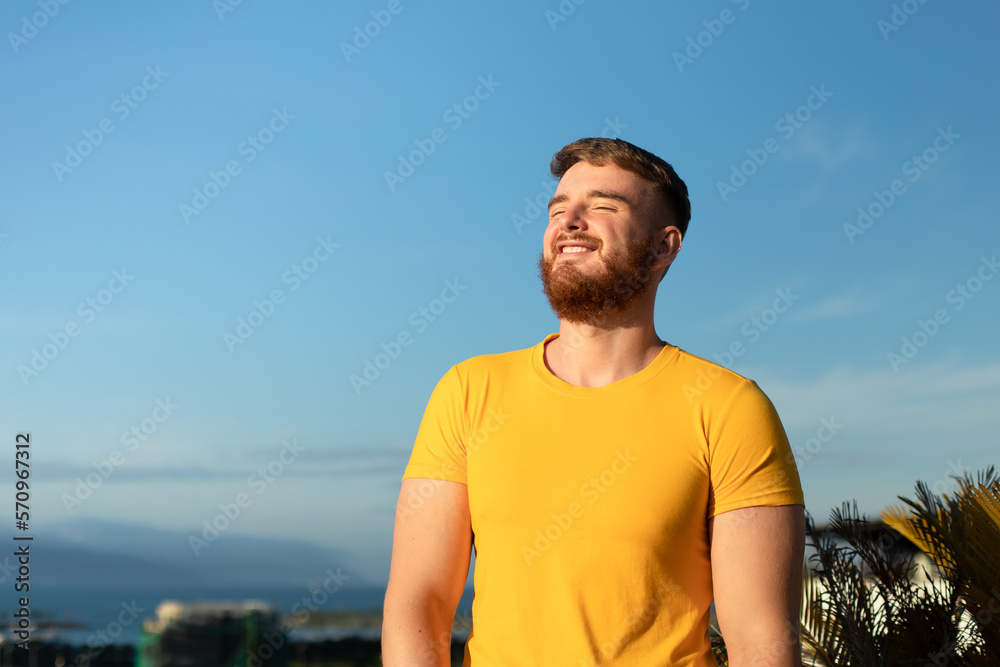 Portrait of happy cheerful positive guy, young handsome bearded man with beard is enjoying sunny day, summer vacation, sunbathing, tanning at sun and smiling, breathing deep, inhale fresh air