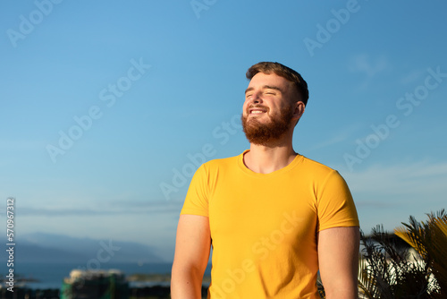 Portrait of happy cheerful positive guy, young handsome bearded man with beard is enjoying sunny day, summer vacation, sunbathing, tanning at sun and smiling, breathing deep, inhale fresh air