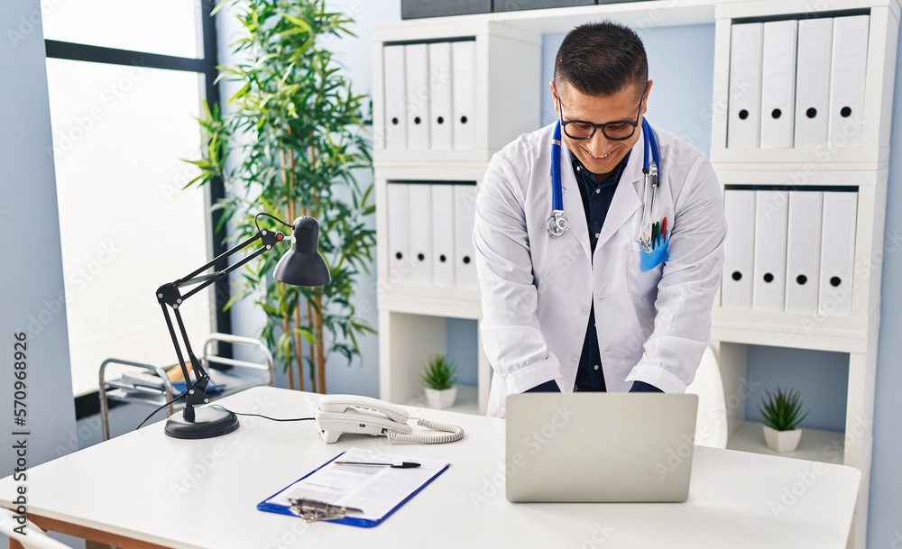 Young latin man doctor using laptop standing at clinic