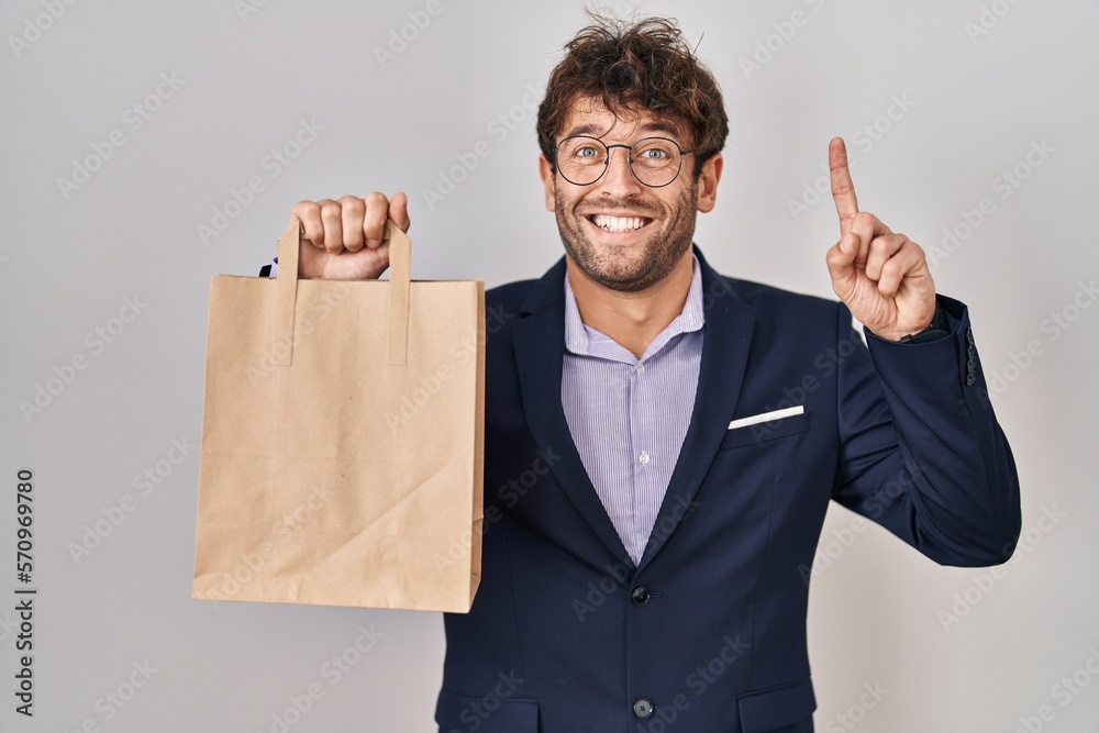 Hispanic business man holding delivery bag surprised with an idea or question pointing finger with happy face, number one Hispanic business man holding delivery bag surprised with an idea or question pointing finger with happy face, number one