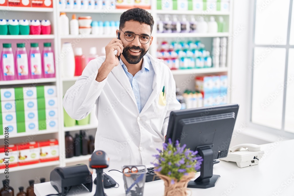 Young arab man pharmacist talking on smartphone using computer at pharmacy