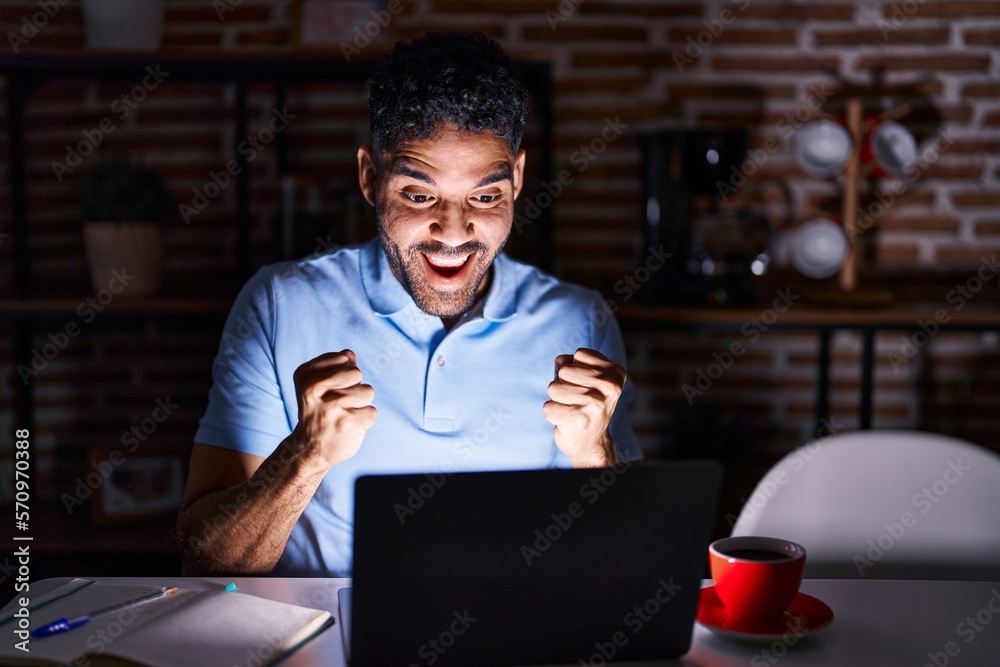 Hispanic man with beard using laptop at night celebrating surprised and amazed for success with arms raised and open eyes. winner concept. Hispanic man with beard using laptop at night celebrating surprised and amazed for success with arms raised and open eyes. winner concept.