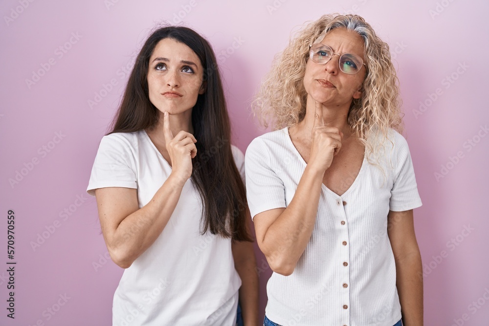 Mother and daughter standing together over pink background thinking concentrated about doubt with finger on chin and looking up wondering Mother and daughter standing together over pink background thinking concentrated about doubt with finger on chin and looking up wondering