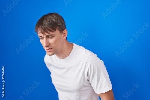 Caucasian blond man standing over blue background suffering of backache, touching back with hand, muscular pain