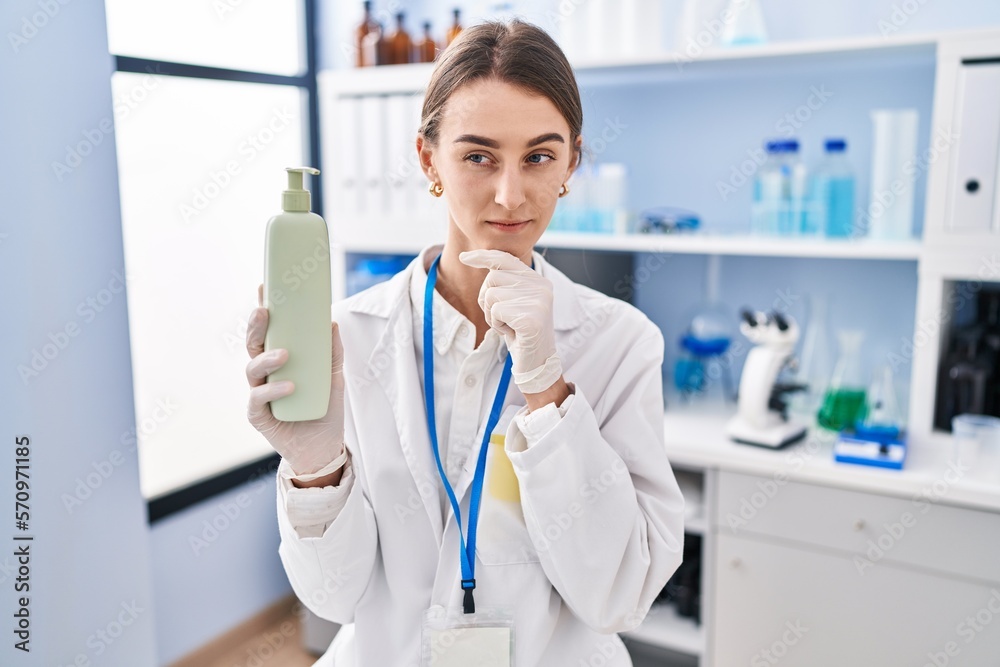 Young caucasian woman working at scientist laboratory holding body lotion serious face thinking about question with hand on chin, thoughtful about confusing idea Young caucasian woman working at scientist laboratory holding body lotion serious face thinking about question with hand on chin, thoughtful about confusing idea
