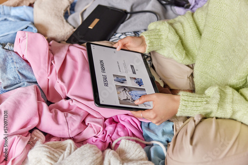 Cropped shot of unrecognizable woman holds tablet surfs resale fashion sites sells clothing online wears jumper and trousers. View from above. Selling something secondhand. E commerce concept