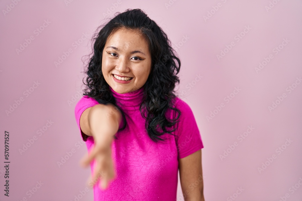 Young asian woman standing over pink background smiling friendly offering handshake as greeting and welcoming. successful business. Young asian woman standing over pink background smiling friendly offering handshake as greeting and welcoming. successful business.