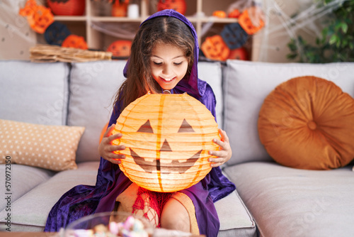 Adorable hispanic girl wearing halloween costume holding pumpkin basket lamp at home