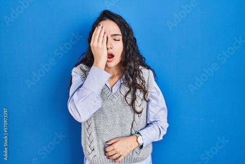 Young brunette woman standing over blue background yawning tired covering half face, eye and mouth with hand. face hurts in pain.