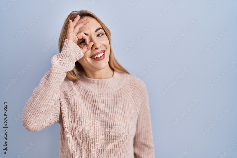 Hispanic woman standing over blue background doing ok gesture with hand smiling, eye looking through fingers with happy face. Hispanic woman standing over blue background doing ok gesture with hand smiling, eye looking through fingers with happy face.