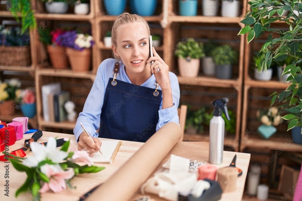 Young blonde woman florist talking on smartphone writing on notebook at florist Young blonde woman florist talking on smartphone writing on notebook at florist