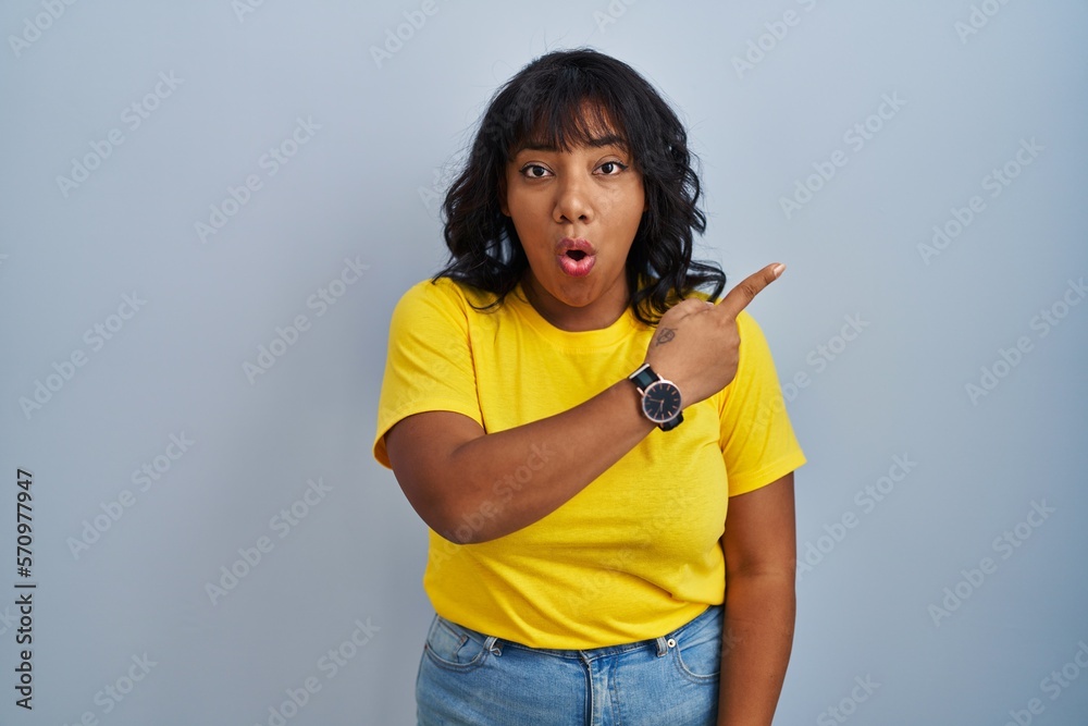 Hispanic woman standing over blue background surprised pointing with finger to the side, open mouth amazed expression. Hispanic woman standing over blue background surprised pointing with finger to the side, open mouth amazed expression.