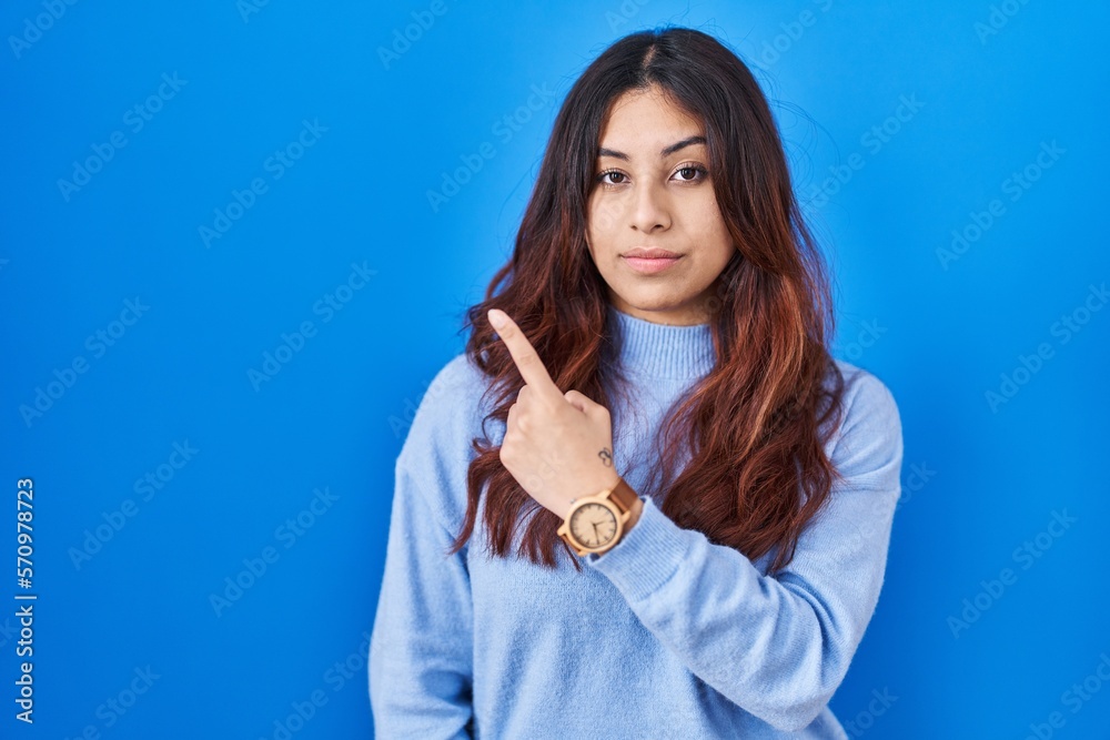 Hispanic young woman standing over blue background pointing with hand finger to the side showing advertisement, serious and calm face Hispanic young woman standing over blue background pointing with hand finger to the side showing advertisement, serious and calm face