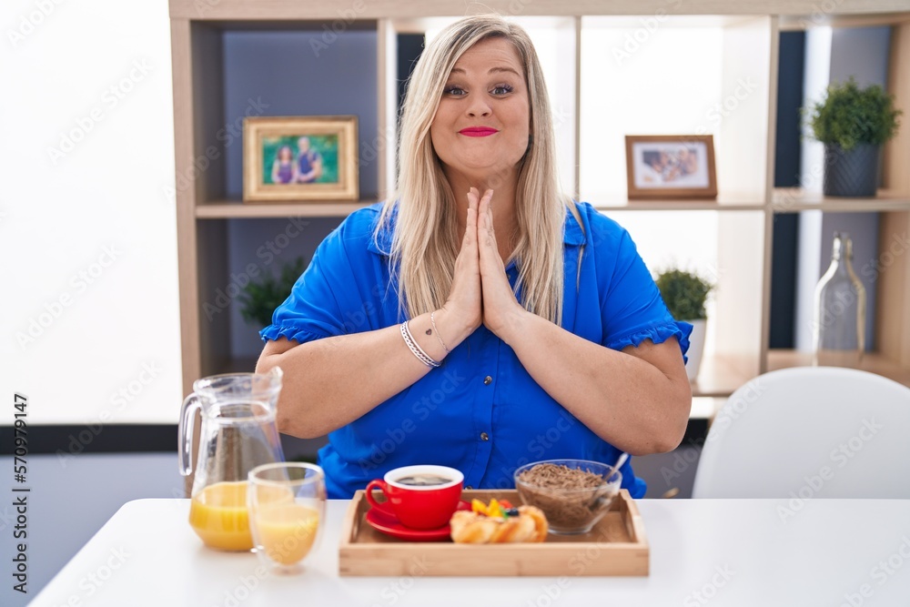 Caucasian plus size woman eating breakfast at home praying with hands together asking for forgiveness smiling confident.