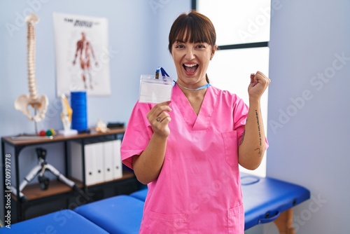 Young brunette woman working at rehabilitation clinic showing id screaming proud  celebrating victory and success very excited with raised arm