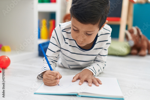 Adorable hispanic boy student writing on notebook at kindergarten