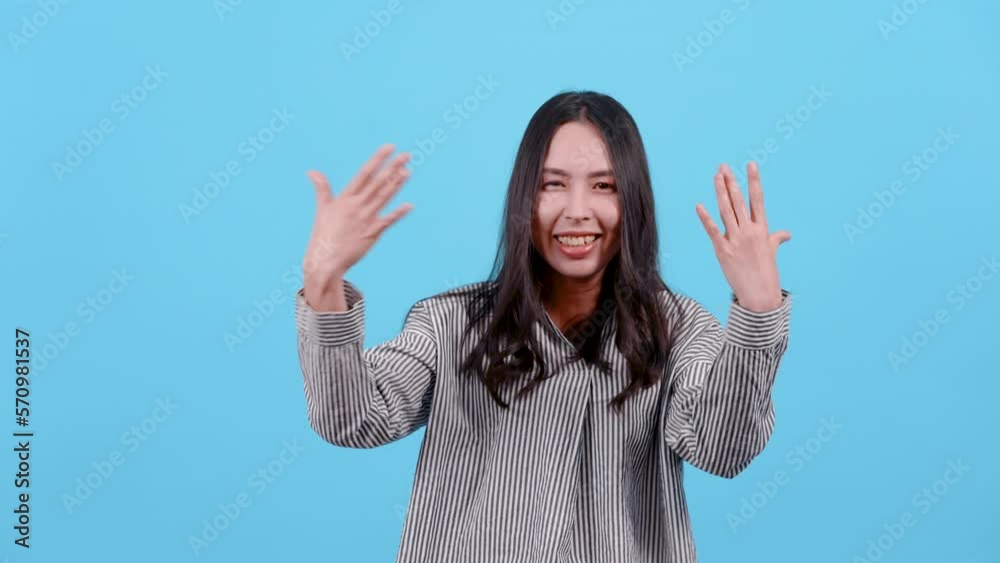 4K, Asian woman with long hair wearing black white striped shirt, raised both hands high, flicked toward himself, showing signs of joy, Isolated indoor studio on blue background.