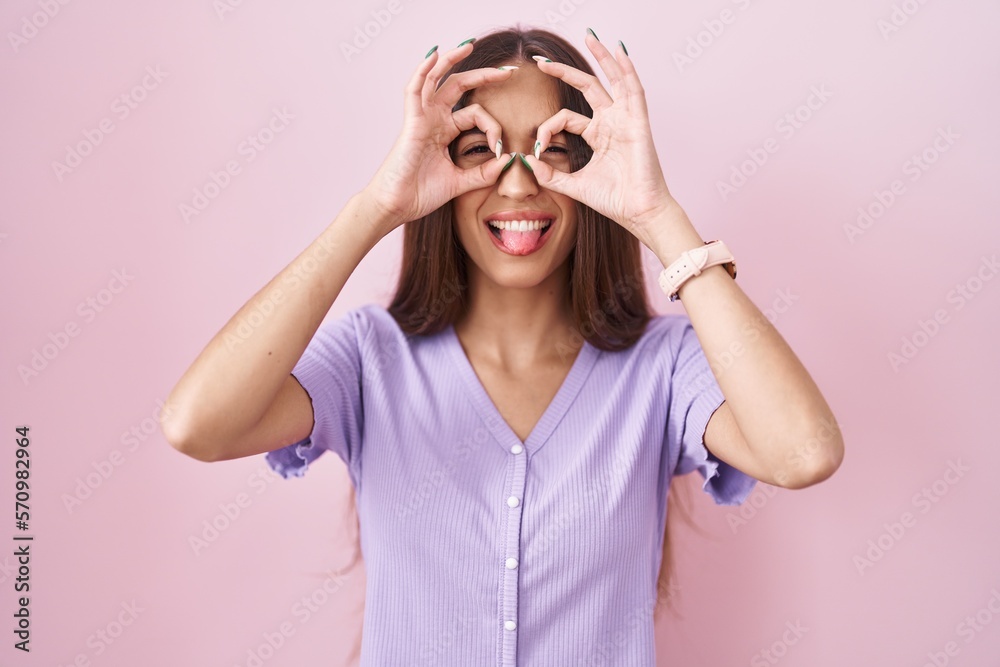 Young hispanic woman with long hair standing over pink background doing ok gesture like binoculars sticking tongue out, eyes looking through fingers. crazy expression. Young hispanic woman with long hair standing over pink background doing ok gesture like binoculars sticking tongue out, eyes looking through fingers. crazy expression.
