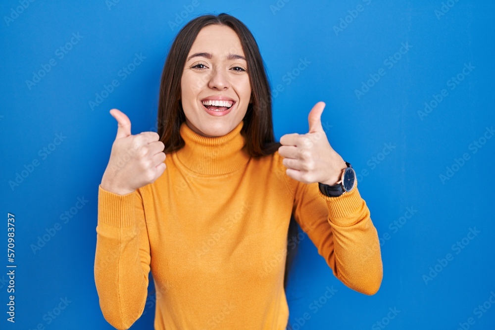 Young brunette woman standing over blue background success sign doing positive gesture with hand, thumbs up smiling and happy. cheerful expression and winner gesture. Young brunette woman standing over blue background success sign doing positive gesture with hand, thumbs up smiling and happy. cheerful expression and winner gesture.