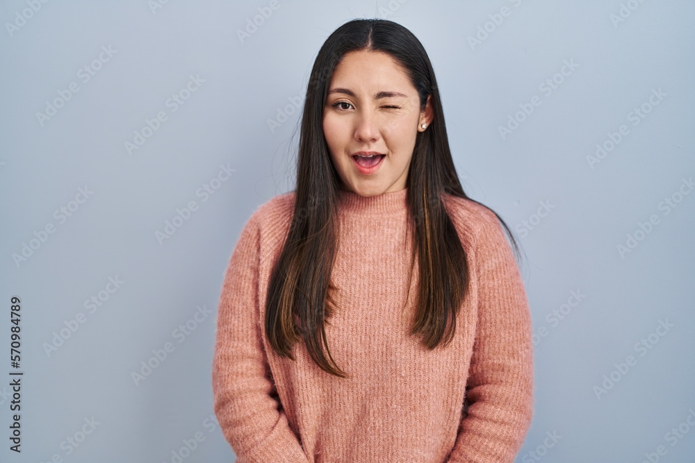 Fototapeta premium Young latin woman standing over blue background winking looking at the camera with sexy expression, cheerful and happy face.