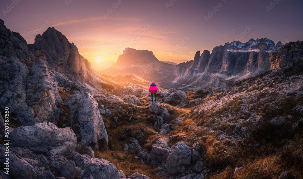 Fototapeta premium Woman on the mountain trail and stones at sunset in autumn. Hiking in Dolomites, Italy. Girl on the path and high rocks at twilight in fall. Colorful landscape with cliffs, grass, golden sky. Nature