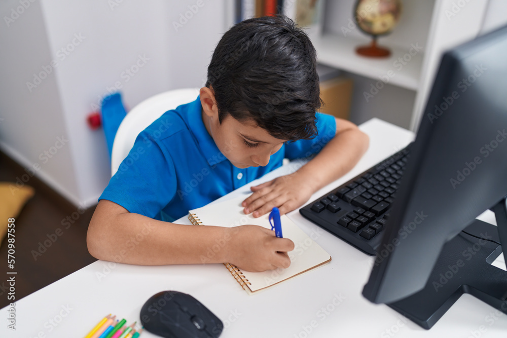 Adorable hispanic boy student using computer writing on notebook at classroom Adorable hispanic boy student using computer writing on notebook at classroom