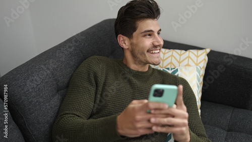 Young hispanic man using smartphone lying on sofa at home