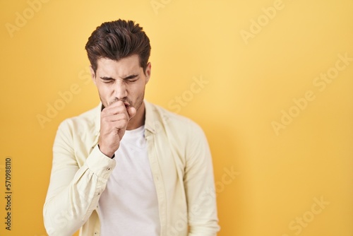Young hispanic man standing over yellow background feeling unwell and coughing as symptom for cold or bronchitis. health care concept.