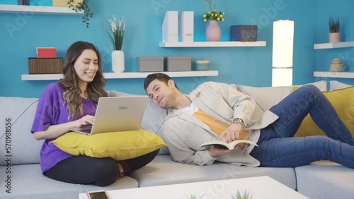 Happy couple sitting on sofa at home having a pleasant and intimate time.
Happy couple having fun together at home. The man reading a book is looking at his wife working on the laptop, they are chatti photo