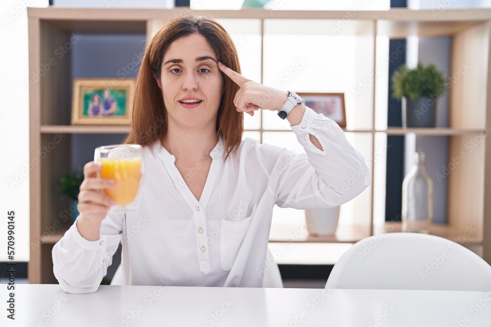 Brunette woman drinking glass of orange juice smiling pointing to head with one finger, great idea or thought, good memory Brunette woman drinking glass of orange juice smiling pointing to head with one finger, great idea or thought, good memory