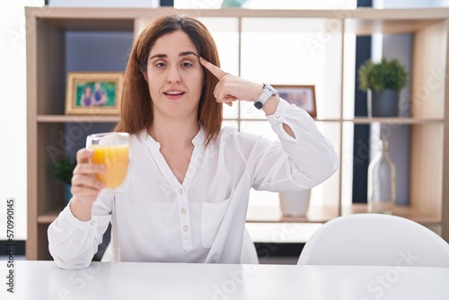 Brunette woman drinking glass of orange juice smiling pointing to head with one finger  great idea or thought  good memory