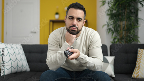 Young hispanic man watching tv sitting on sofa with boring expression at home