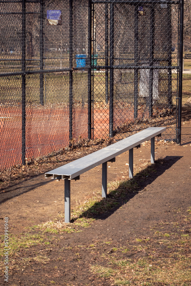 Close up of an empty aluminum metal baseball bench in a grass and dirt dugout with black chain link fence and muddy red colored infield beyond. Close up of an empty aluminum metal baseball bench in a grass and dirt dugout with black chain link fence and muddy red colored infield beyond.