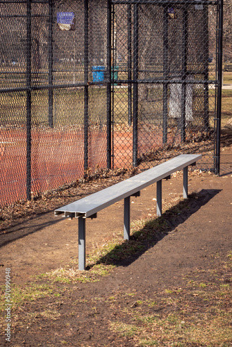 Close up of an empty aluminum metal baseball bench in a grass and dirt dugout with black chain link fence and muddy red colored infield beyond.