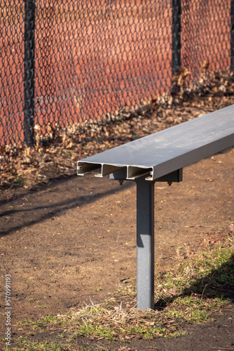 Close up of an empty aluminum metal baseball bench in a grass and dirt dugout with black chain link fence and muddy red colored infield beyond. © Joseph Kirsch