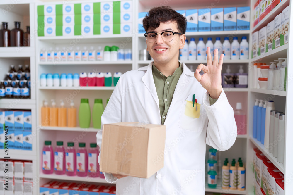 Young non binary man working at pharmacy drugstore holding box doing ok sign with fingers, smiling friendly gesturing excellent symbol Young non binary man working at pharmacy drugstore holding box doing ok sign with fingers, smiling friendly gesturing excellent symbol