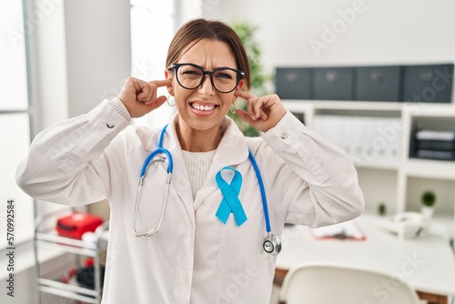 Young brunette doctor woman wearing stethoscope at the clinic covering ears with fingers with annoyed expression for the noise of loud music. deaf concept.