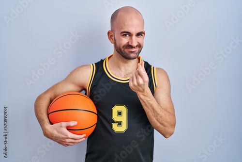 Young bald man with beard wearing basketball uniform holding ball doing money gesture with hands, asking for salary payment, millionaire business