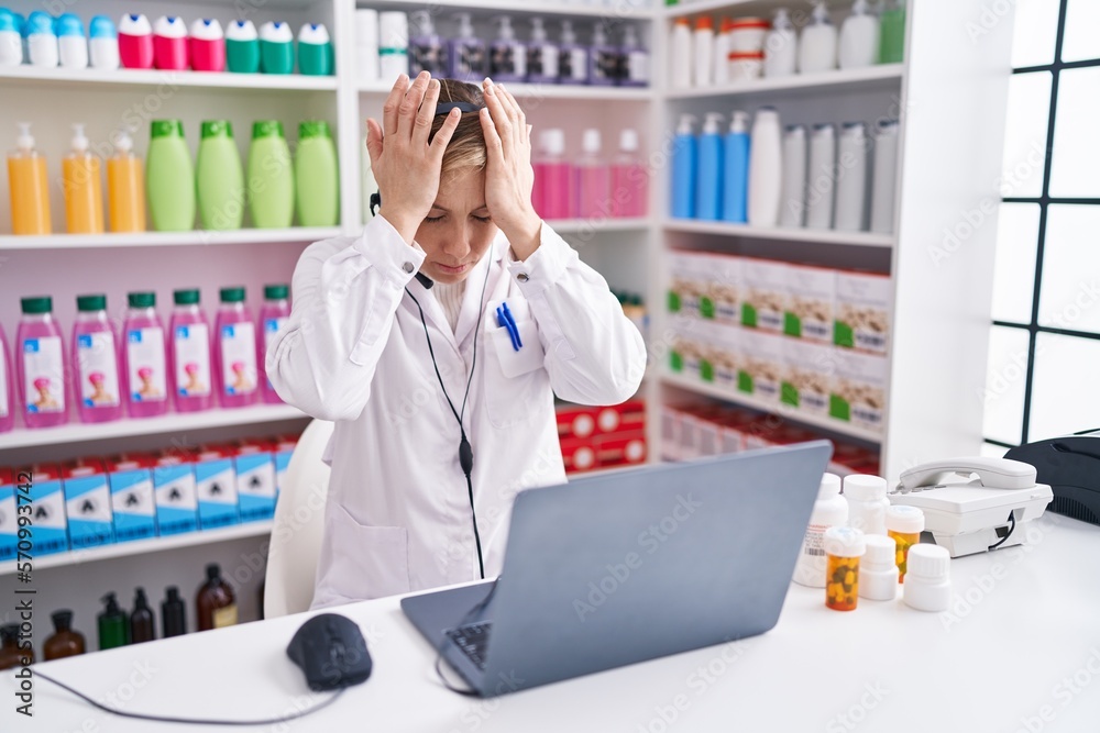 Young caucasian woman working at pharmacy drugstore using laptop suffering from headache desperate and stressed because pain and migraine. hands on head.
