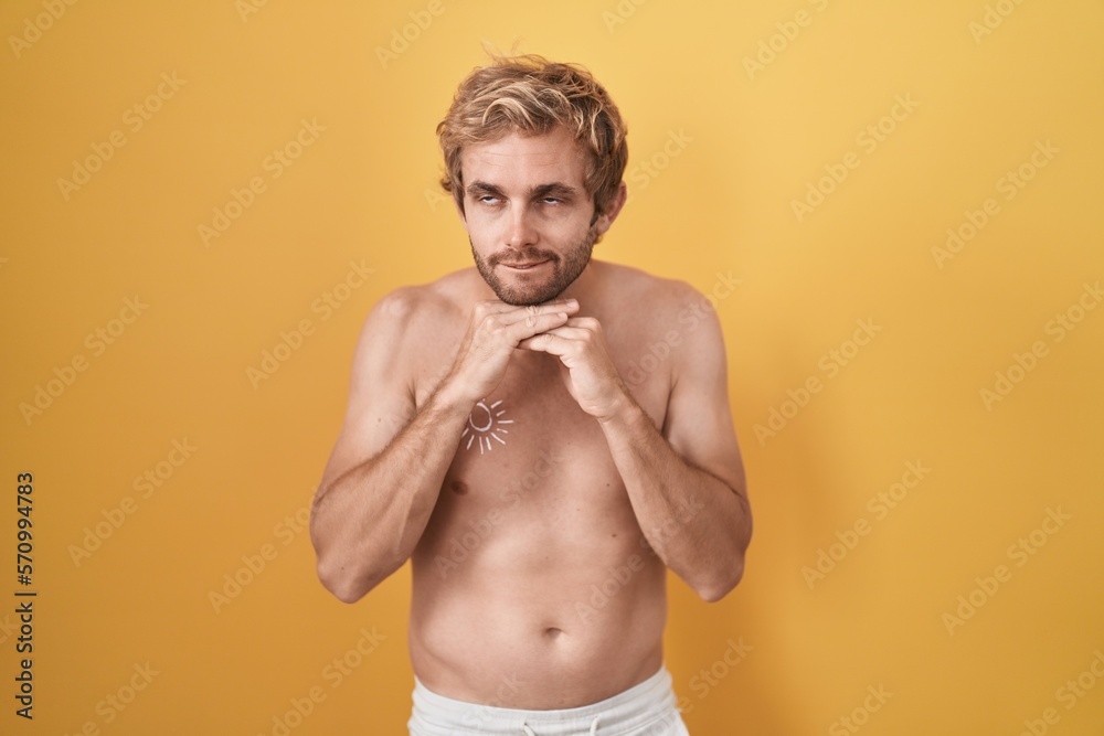 Caucasian man standing shirtless wearing sun screen laughing nervous and excited with hands on chin looking to the side