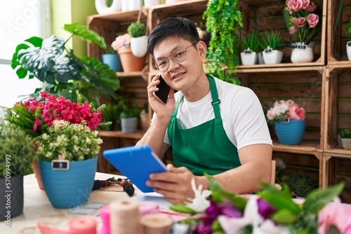 Young chinese man florist talking on smartphone using touchpad at flower shop
