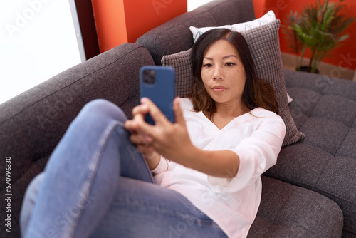 Young asian woman make selfie by smartphone lying on sofa at home