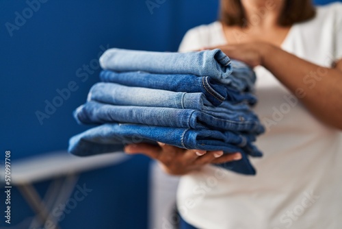 Middle age hispanic woman holding folded jeans at laundry room