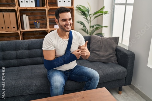 Young hispanic man with beard wearing arm on sling sitting at therapy consult smiling happy and positive, thumb up doing excellent and approval sign