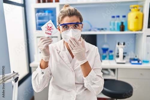 Young woman working at scientist laboratory holding toxic banner covering mouth with hand, shocked and afraid for mistake. surprised expression