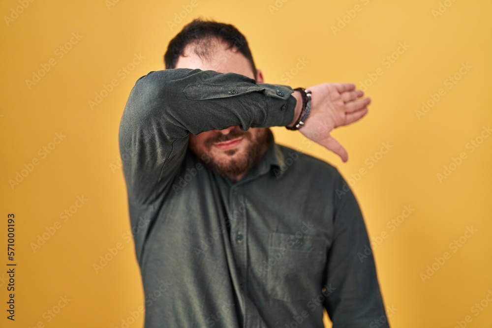 Plus size hispanic man with beard standing over yellow background covering eyes with arm, looking serious and sad. sightless, hiding and rejection concept Plus size hispanic man with beard standing over yellow background covering eyes with arm, looking serious and sad. sightless, hiding and rejection concept
