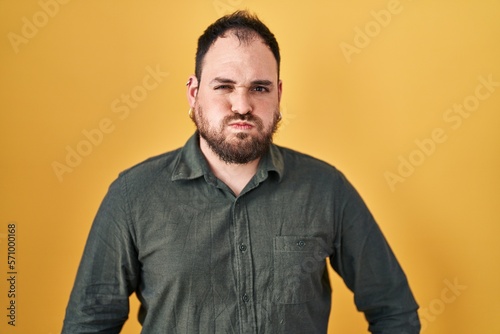 Plus size hispanic man with beard standing over yellow background puffing cheeks with funny face. mouth inflated with air, crazy expression.